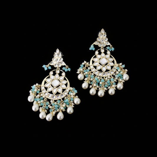 A pair of gold-plated Indian chandelier earrings with white faux pearls, turquoise blue beads, and clear rhinestones, displayed on a dark reflective surface with the SAHELI MY BEST FRIEIND logo at the top.