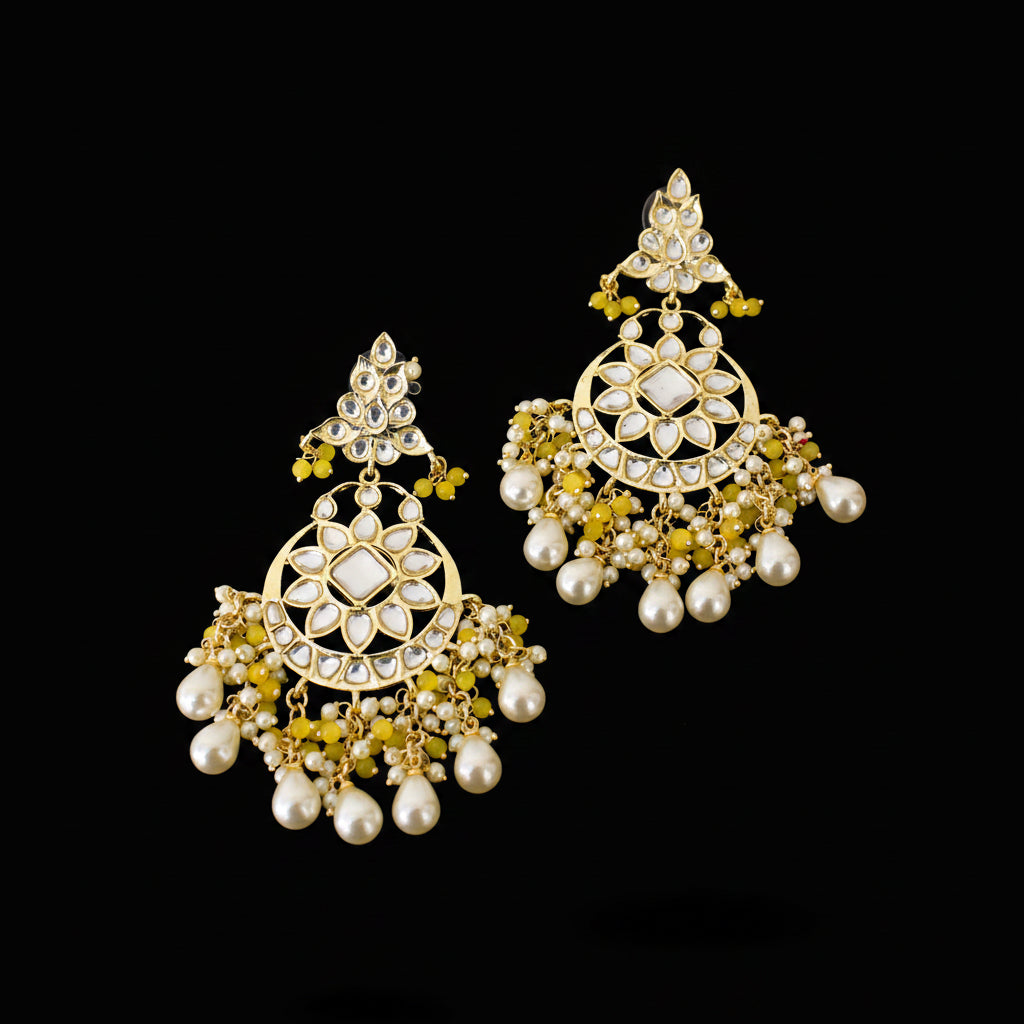 A pair of ornate gold-toned Indian jhumka earrings decorated with white faux pearls, clear crystals, and small olive green floral accents, displayed on a dark reflective surface with the SAHELI brand logo at the top.
