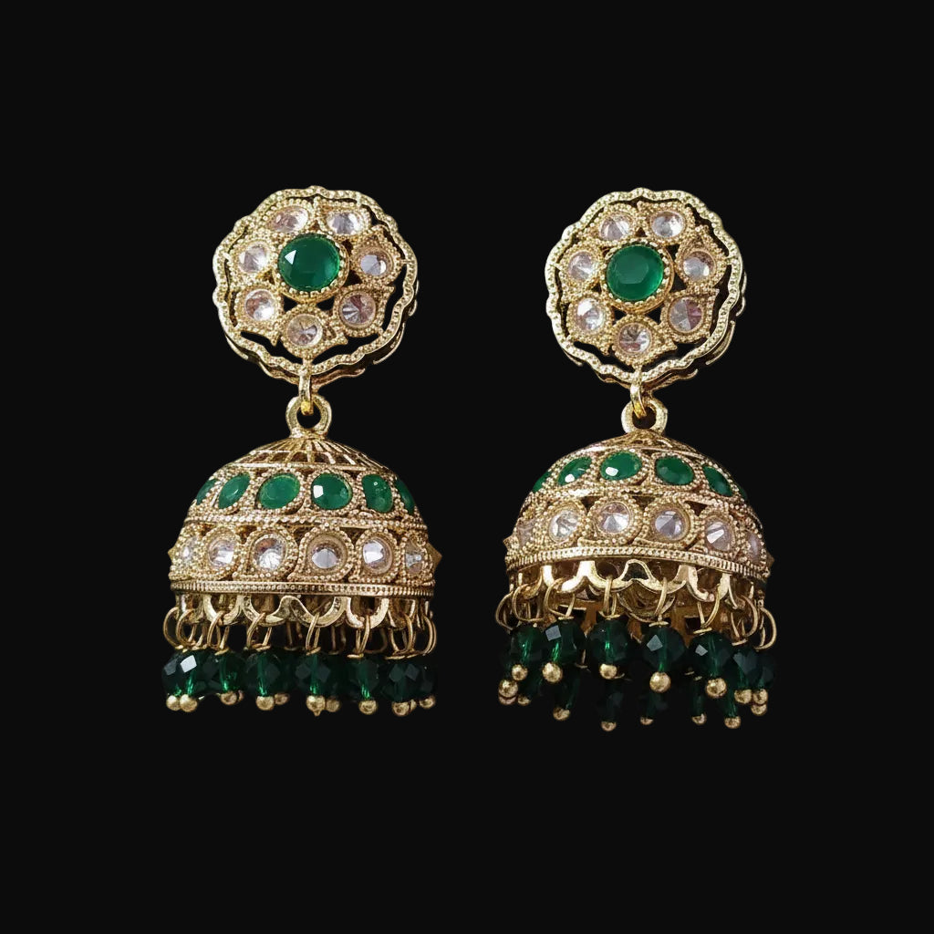 A pair of gold-plated Indian jhumka earrings with green stones and clear rhinestones, displayed on a solid dark pink background.