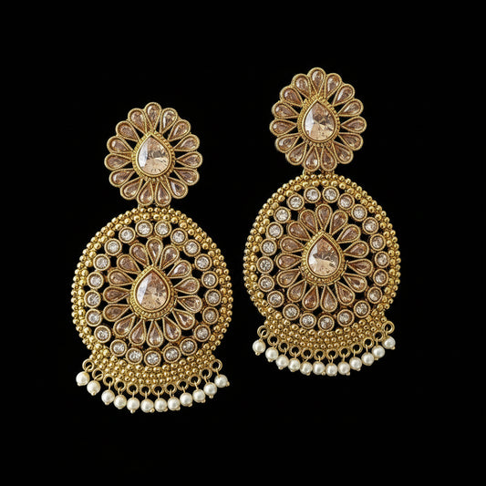 A pair of ornate gold-toned Indian jhumka earrings with intricate beadwork, clear stones, and small white pearl-like dangling beads, displayed on a dark gray background. The brand logo 'SAHELI MY BEST FRIEND' is visible in the top left corner.
