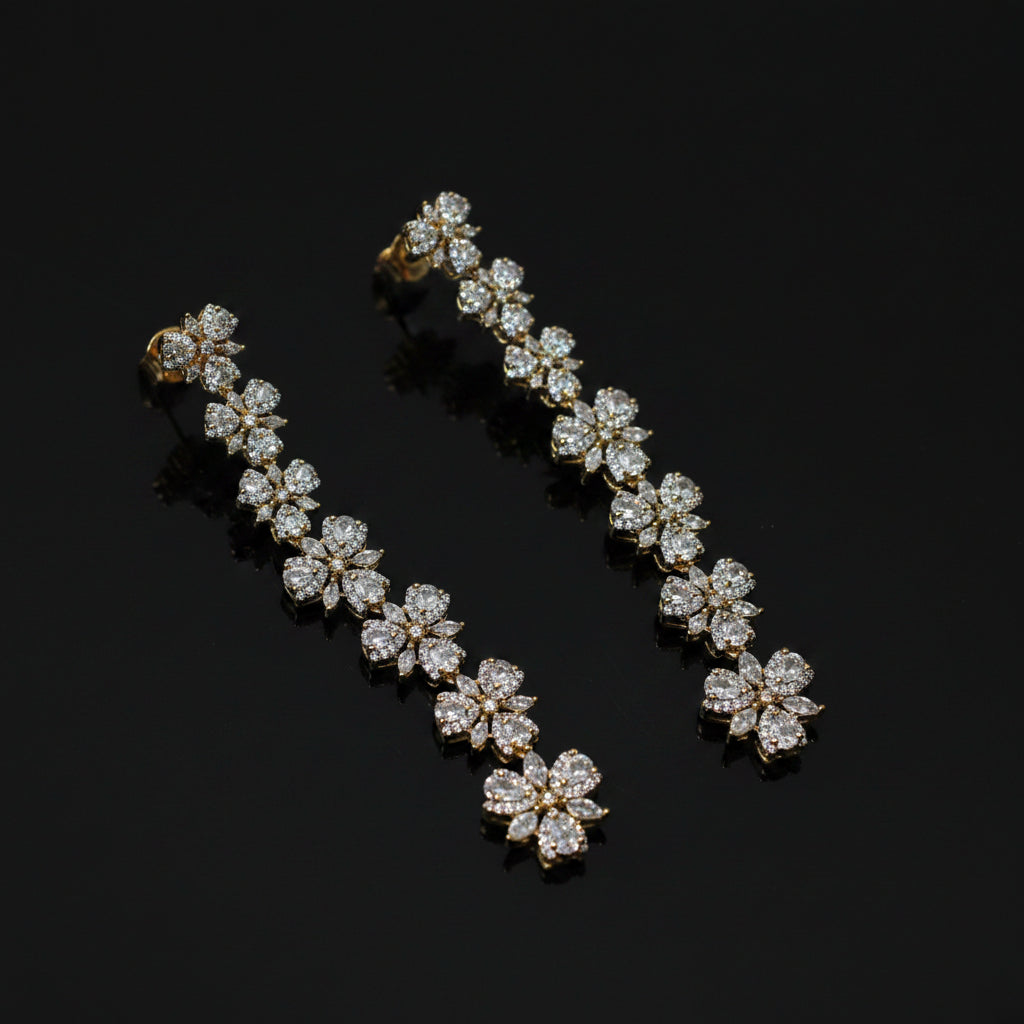 A pair of gold-toned drop earrings with a continuous row of flower-shaped clusters of clear stones, displayed on a dark gray background.
