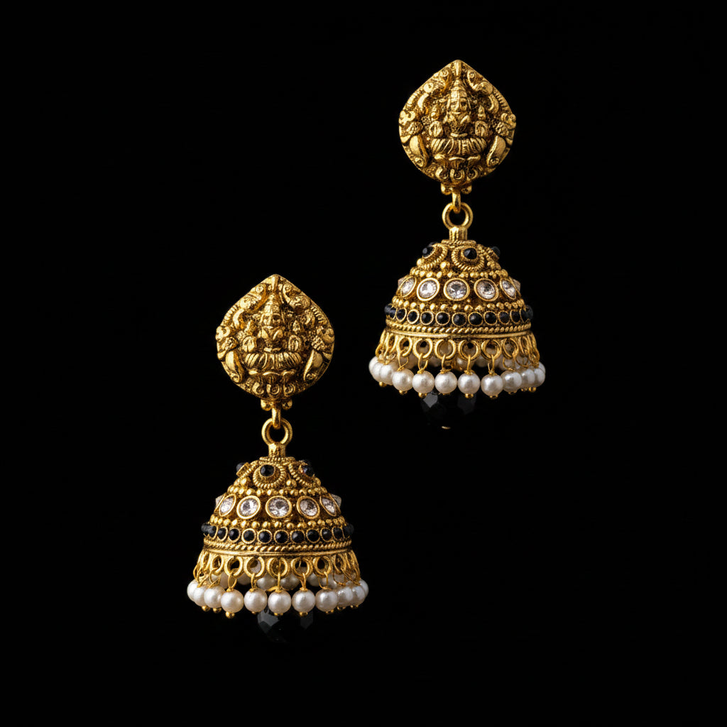 A pair of ornate gold-toned jhumka earrings displayed on a black Y-shaped jewelry stand against a gray gradient background. The earrings feature intricate filigree, black and white stone accents, and dangling pearl bead strands.