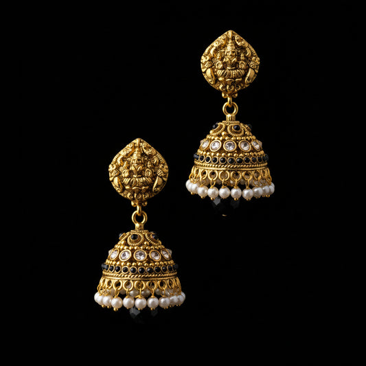 A pair of ornate gold-toned jhumka earrings displayed on a black Y-shaped jewelry stand against a gray gradient background. The earrings feature intricate filigree, black and white stone accents, and dangling pearl bead strands.