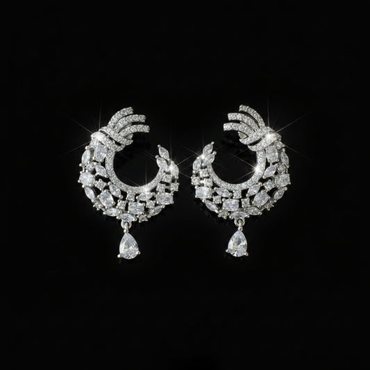 A pair of silver-toned crescent-shaped earrings adorned with clear rhinestones and teardrop dangling stones, displayed on a dark reflective surface with a white SAHeli brand tag.