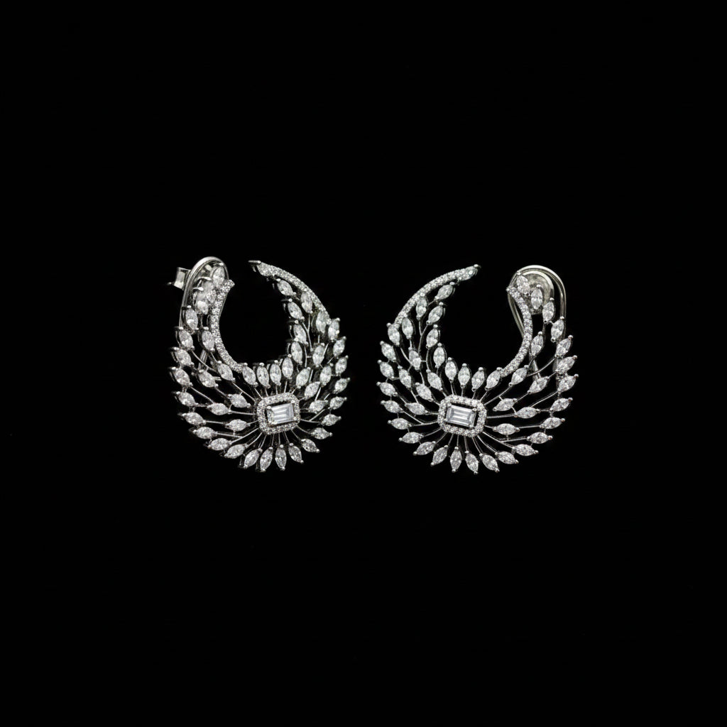 A pair of silver-toned crescent-shaped earrings adorned with numerous clear stones, featuring a central square-cut stone, displayed on a dark gray background.