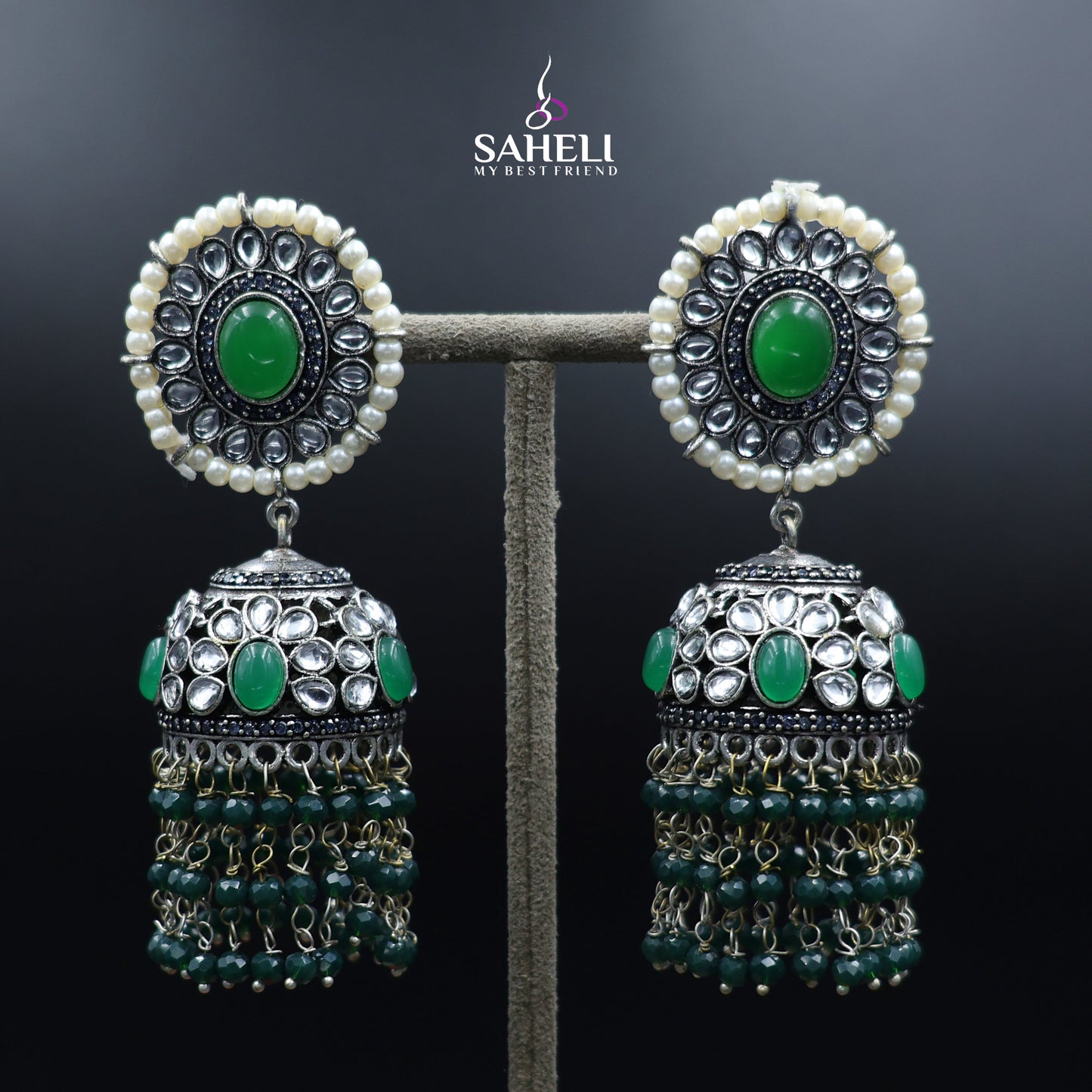 Pair of earrings with green gemstones and pearl-like beads on a dark background, branded 'Sahell'.