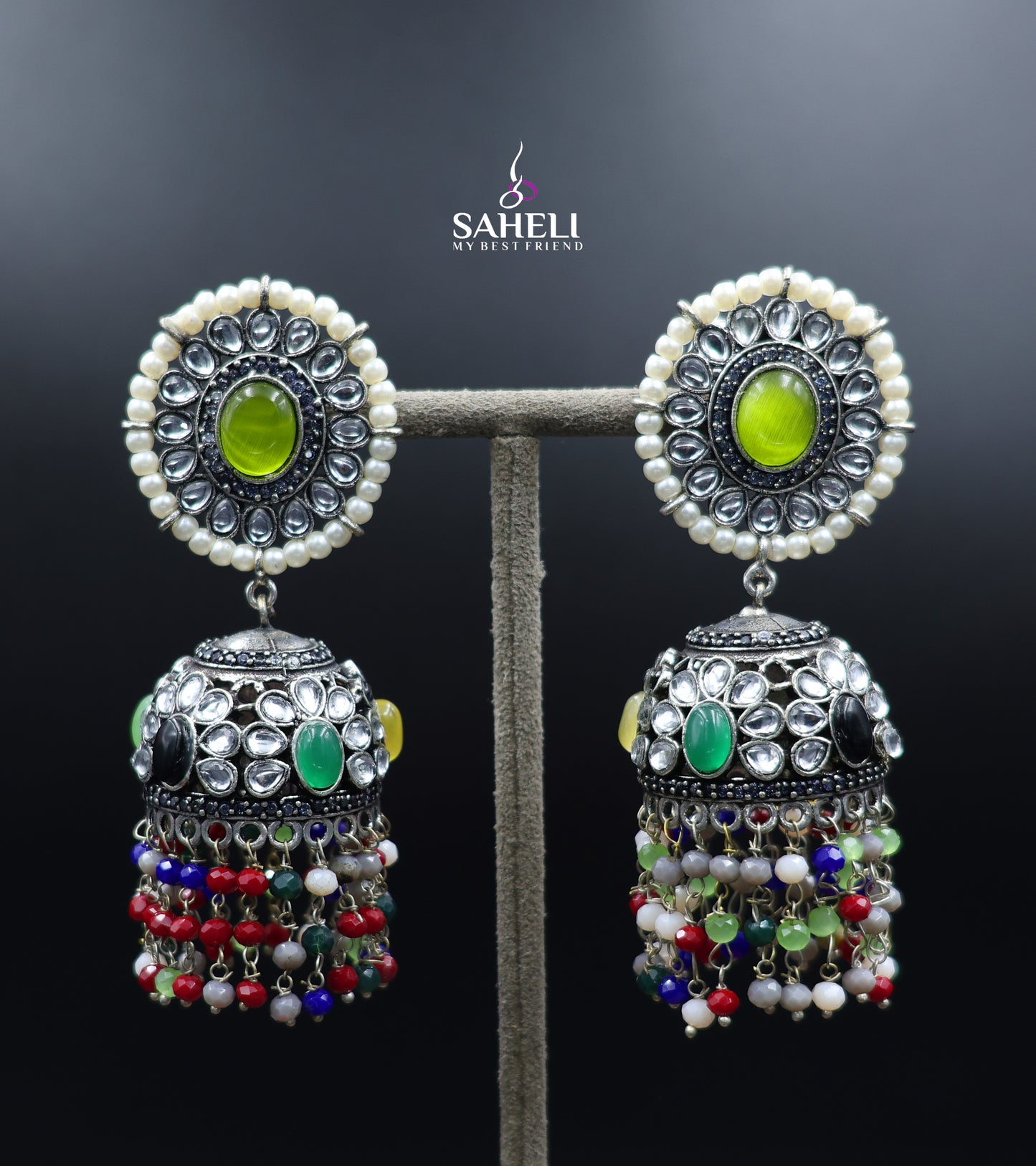 Decorative earrings with green gemstones and colorful beads on a dark background, featuring the brand 'Saheli'.