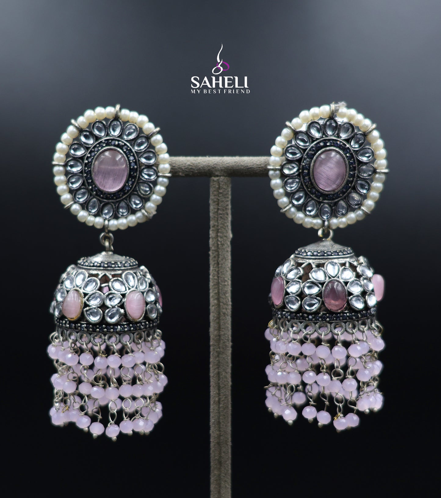 Ornate earrings with pink stones and pearl-like beads on a dark background, branded 'Saheli'.