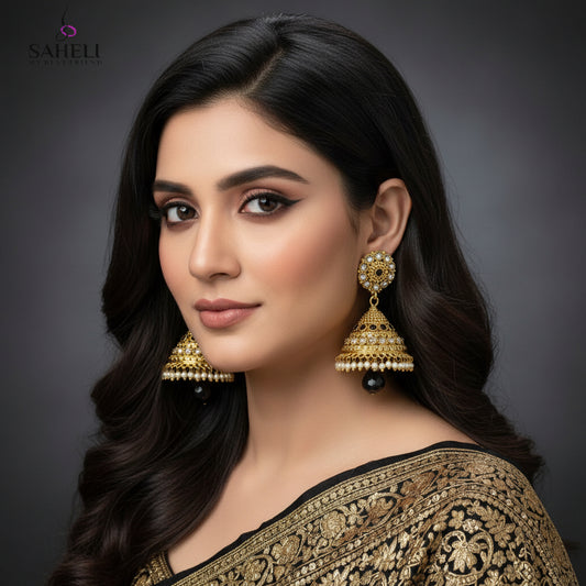 Gold jhumka earrings with black and pearl details on a gray stand, branded 'Saheli'.