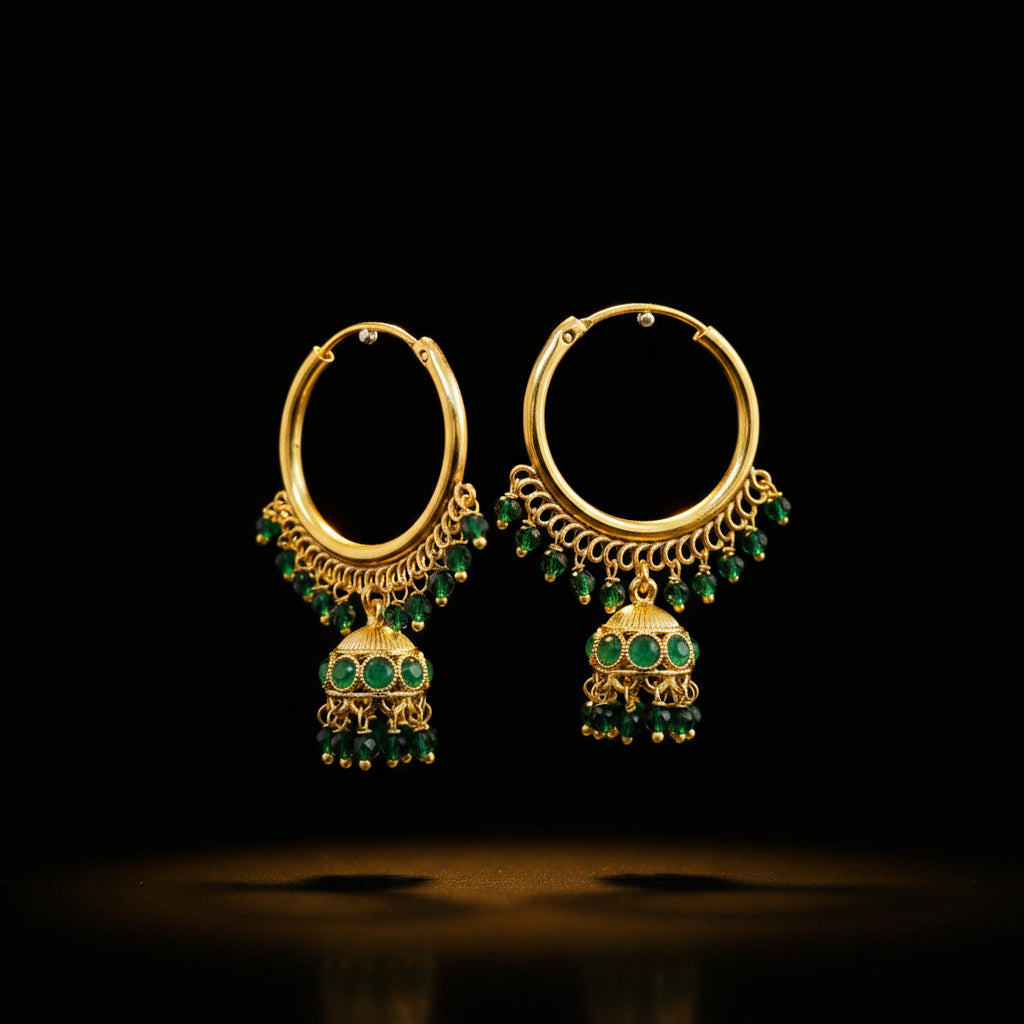 A pair of gold-toned hoop earrings with dangling green stone embellishments and bell-shaped charms, displayed on a gray stand against a gray background. The brand name 'SAHELI MY BEST FRIEND' is printed at the top center.