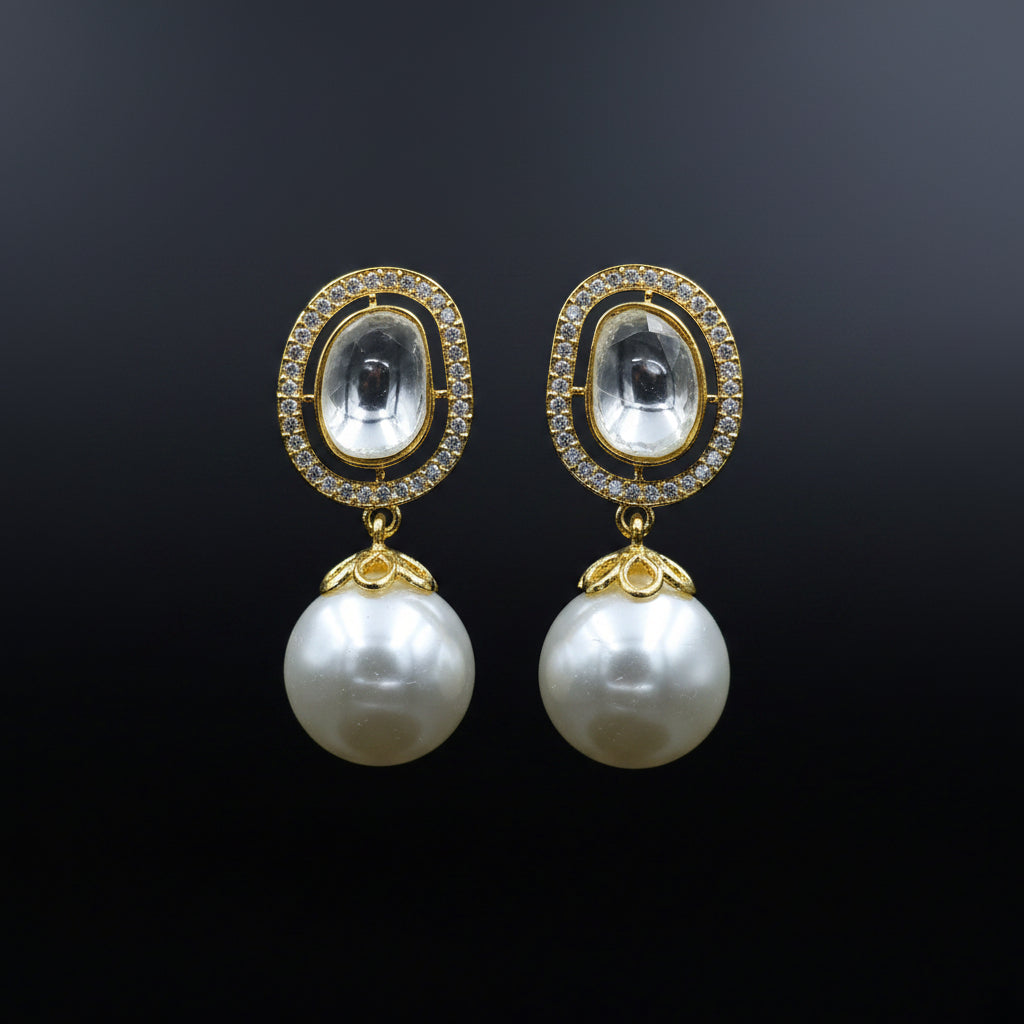 A pair of gold-plated drop earrings with oval clear crystal cabochons surrounded by small clear stones, and a large white pearl drop below, displayed on a black velvet stand against a black background. The brand logo 'SAHELI MYBESTFRIEND' is visible at the top center.