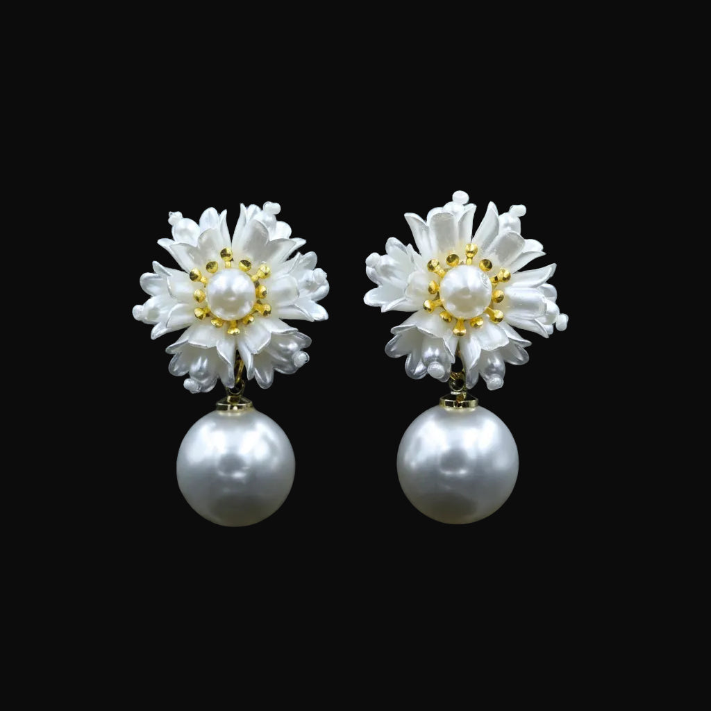 A pair of white flower-shaped drop earrings with pearl centers and gold accents, displayed on a black jewelry stand against a dark background. The brand logo 'SAHELI MY BEST FRIEND' is visible at the top center.
