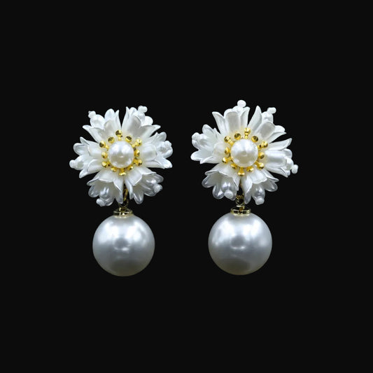 A pair of white flower-shaped drop earrings with pearl centers and gold accents, displayed on a black jewelry stand against a dark background. The brand logo 'SAHELI MY BEST FRIEND' is visible at the top center.