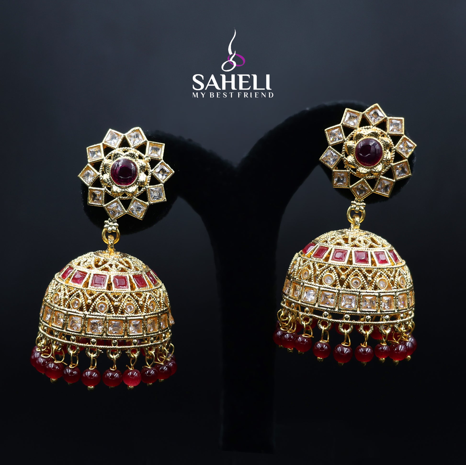 Ornate gold earrings with red gemstones on a dark background, featuring the brand 'Saheli'.