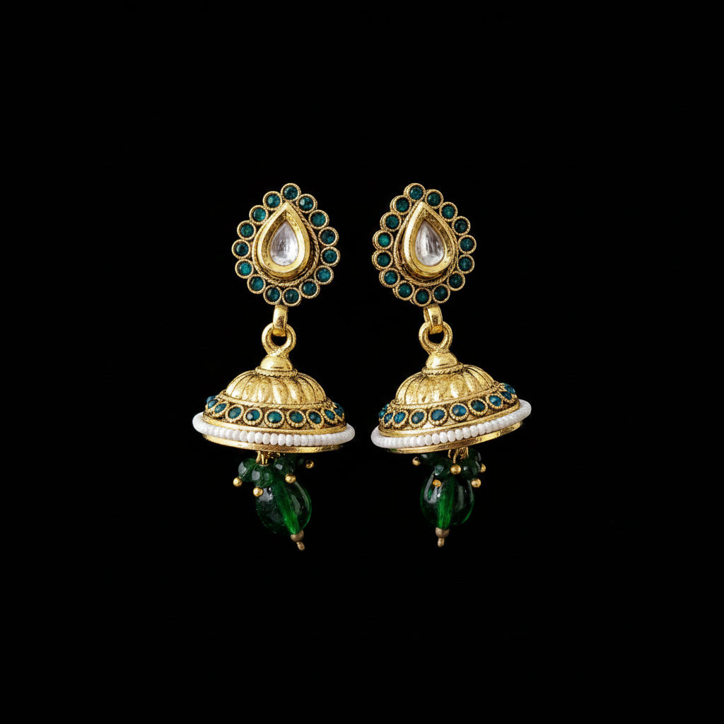 A pair of traditional Indian gold-plated jhumka earrings with green and white stones, displayed on a gray Y-shaped stand against a black background. The brand logo 'SAHELI' is visible at the top center.