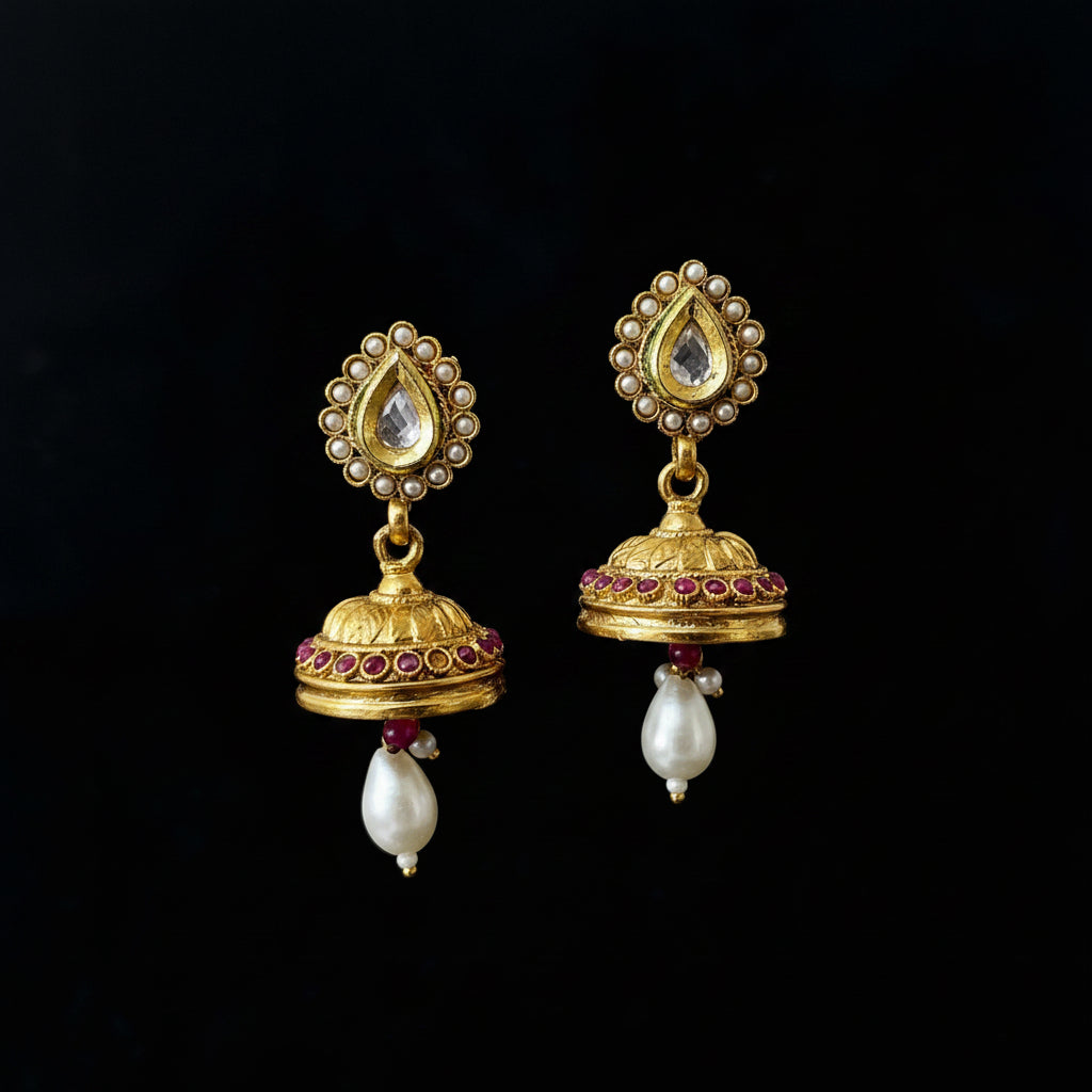 A pair of gold-toned traditional Indian jhumka earrings with white pearls, pink stones, and a central clear teardrop stone, displayed on a black jewelry stand. The brand logo 'SAHELI MY BEST FRIEND' is visible at the top center.