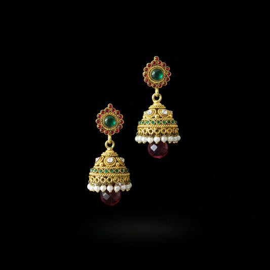 A pair of traditional Indian gold-plated jhumka earrings with green and red stones, white pearl bead borders, and dangling red faceted gems, displayed on a black stand against a dark gray background. The brand logo 'SAHELI' is visible at the top.