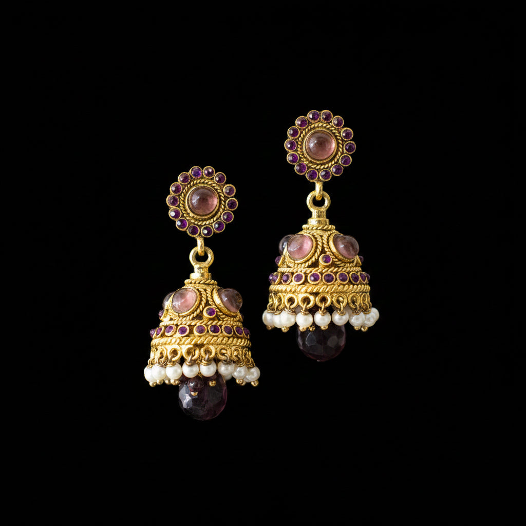 A pair of traditional Indian gold jhumka earrings with purple and white stones, displayed on a black stand against a gray background with the brand logo 'SAHELI MY BESTFRIEND' at the top.