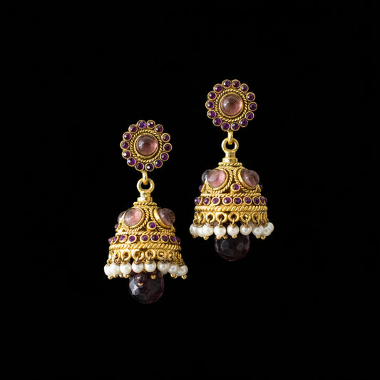 A pair of traditional Indian gold jhumka earrings with purple and white stones, displayed on a black stand against a gray background with the brand logo 'SAHELI MY BESTFRIEND' at the top.