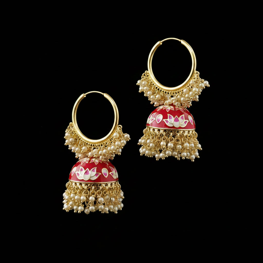 A pair of traditional Indian gold hoop earrings with red and white floral enamel jhumka bells and pearl bead fringe, displayed on a black stand against a gray background.