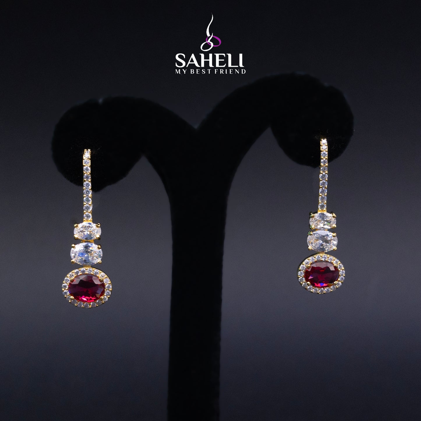 Pair of earrings with gemstones on a black stand, branded 'Saheli'.