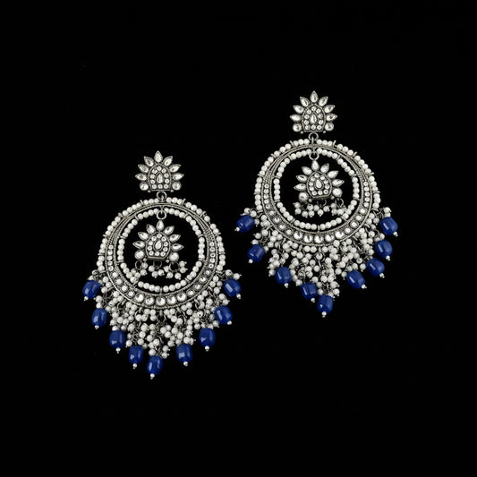 A pair of ornate silver-toned dangle earrings decorated with white beads, clear stones, and blue bead tassels, displayed on a dark reflective surface.