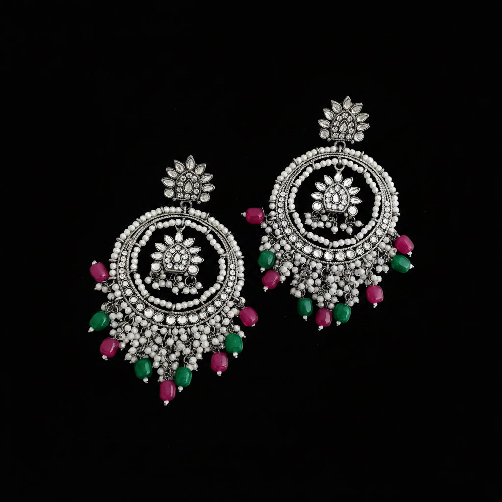 A pair of ornate silver-toned dangle earrings with intricate beadwork and dangling pink and green gemstone accents, displayed on a dark reflective surface.