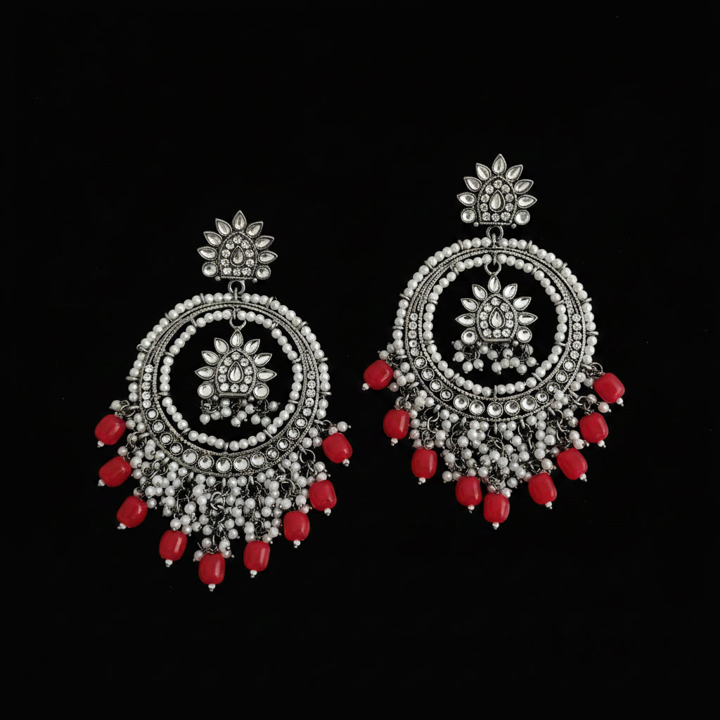 A pair of ornate silver-toned Indian jhumka earrings decorated with clear stones, white beads, and dangling red faceted beads, displayed on a dark reflective surface.