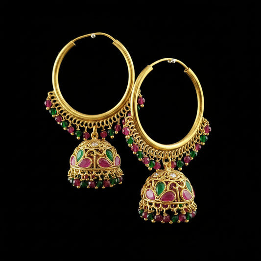A pair of gold-plated Indian jhumka hoop earrings with dangling bell-shaped charms, decorated with pink and green enamel and small red and green stones, displayed on a gray stand.