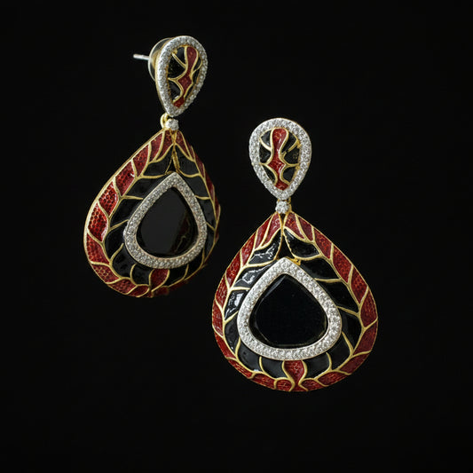 A pair of ornate teardrop-shaped earrings with a black center, red and gold floral patterns, and a silver-tone border set with clear stones, displayed on a dark reflective surface.