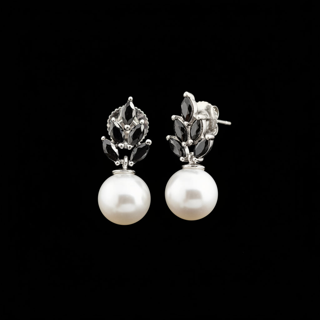A pair of silver-toned drop earrings with white pearl-like round drops and black floral-shaped accents, displayed on a gray jewelry stand against a gray background.