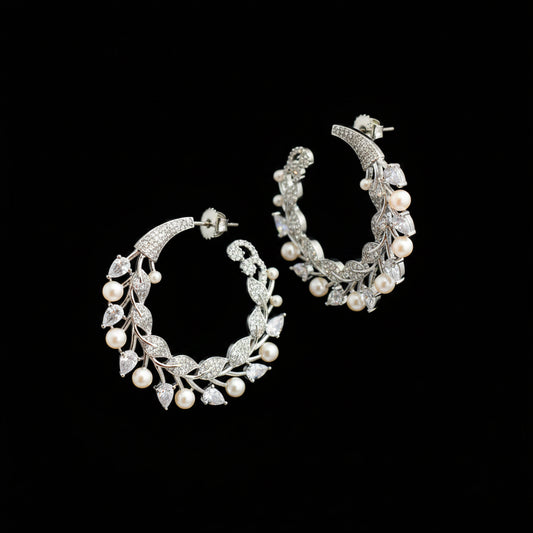 A pair of silver-toned crescent-shaped hoop earrings decorated with leaf and pearl motifs, displayed on a dark reflective surface.