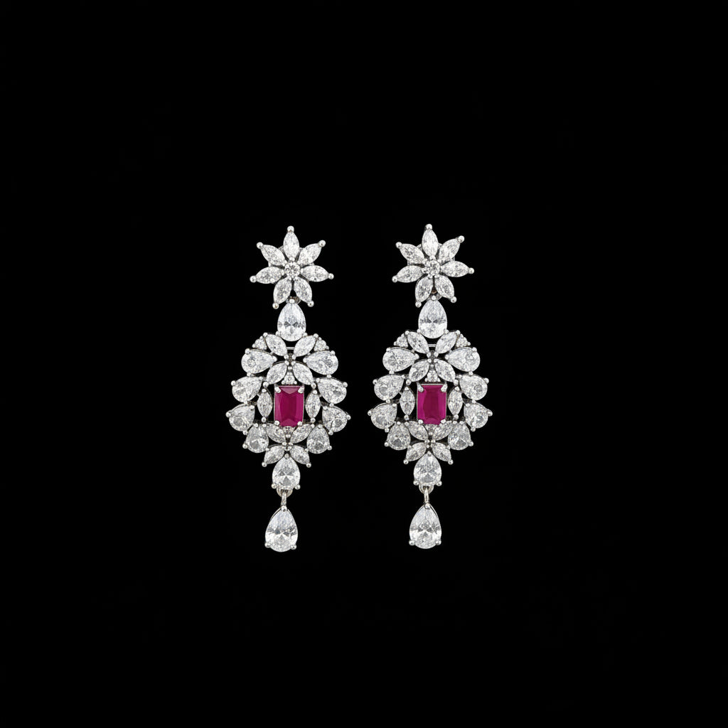 A pair of silver-toned dangle earrings with clear crystal floral motifs and rectangular pink gemstone centers, displayed on a black stand against a dark gray background. The brand logo 'SAHELI' is visible at the top.