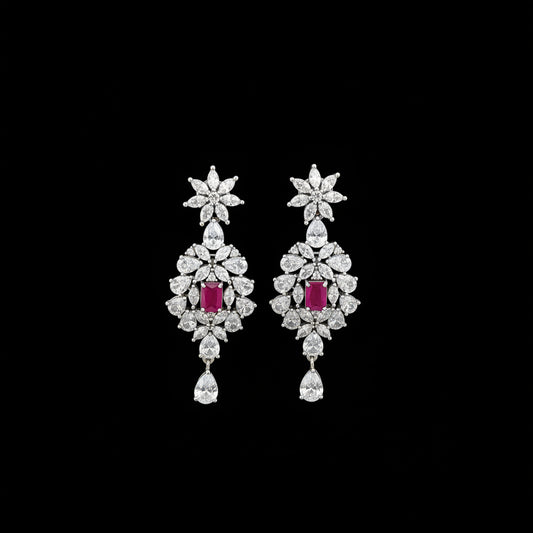 A pair of silver-toned dangle earrings with clear crystal floral motifs and rectangular pink gemstone centers, displayed on a black stand against a dark gray background. The brand logo 'SAHELI' is visible at the top.