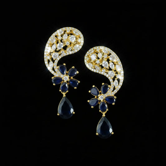 A pair of gold-toned traditional Indian jhumka earrings with deep blue stones and clear pavé accents, displayed on a dark reflective surface with a Saheli by Srinivasand logo in the top left corner.