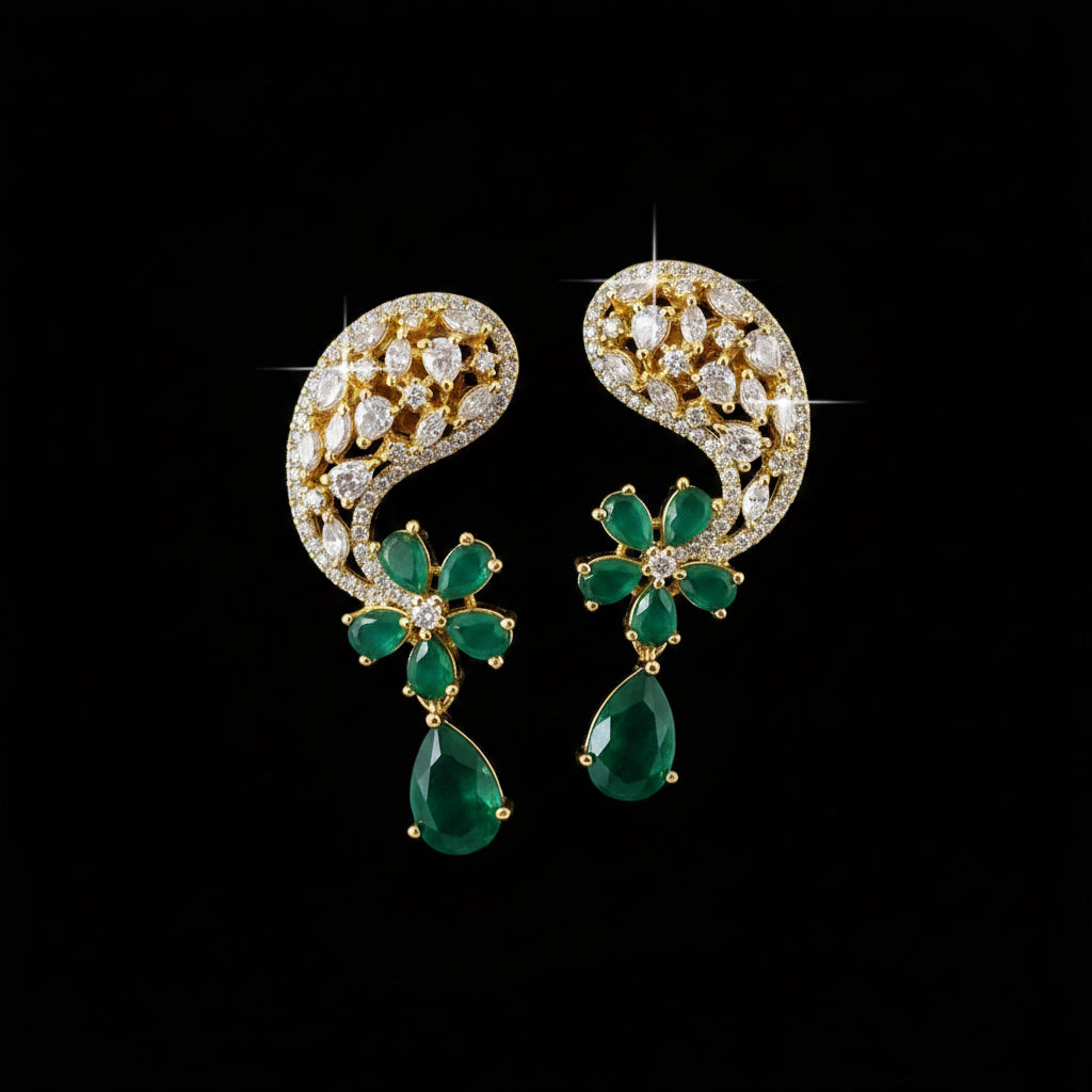 A pair of gold-plated statement earrings featuring green emerald-cut stones and clear pavé-set stones, displayed on a neutral gray background with the 'SAHELI MY BEST FRIEND' logo at the top.