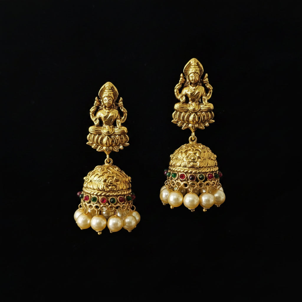 A pair of gold-toned traditional Indian jhumka earrings displayed on a black Y-shaped jewelry stand. Each earring features a deity figure at the top, an ornate bell-shaped dangle, and rows of white faux pearls with small red and green stone accents.