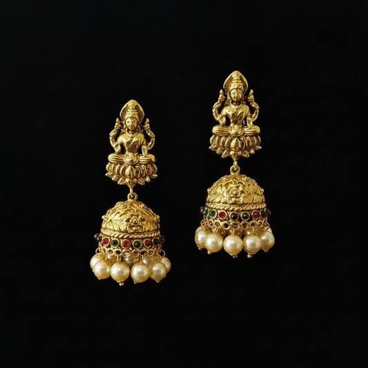 A pair of gold-toned traditional Indian jhumka earrings displayed on a black Y-shaped jewelry stand. Each earring features a deity figure at the top, an ornate bell-shaped dangle, and rows of white faux pearls with small red and green stone accents.