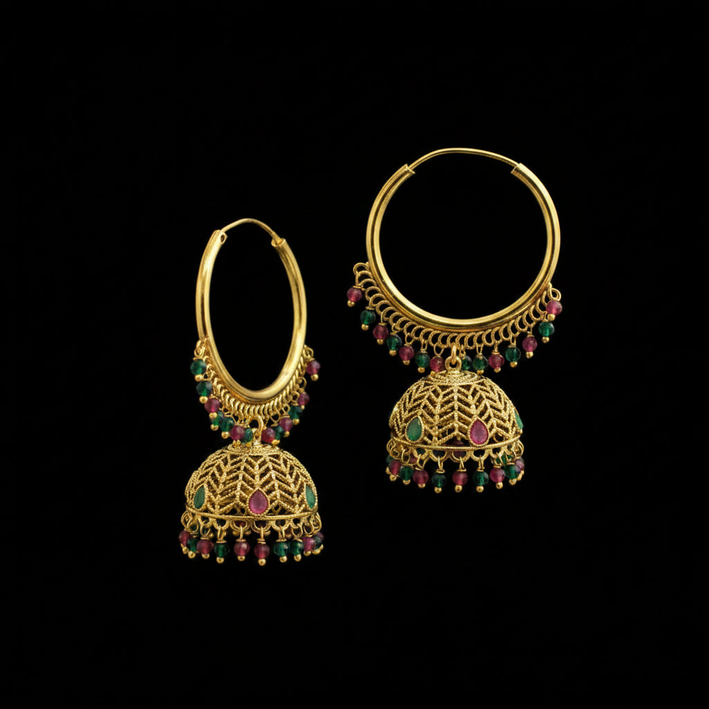 A pair of gold-toned traditional hoop earrings with dangling filigree jhumka bells and small green and pink gemstone accents, displayed on a dark gray stand against a gray background.