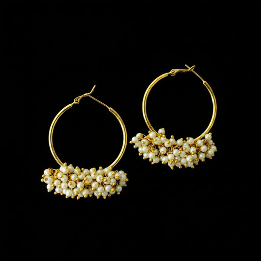 A pair of gold hoop earrings with a cluster of small white pearl-like beads at the bottom, displayed on a dark reflective surface. The brand logo 'SAHeli MY BEST FRIEND' is visible at the top center.
