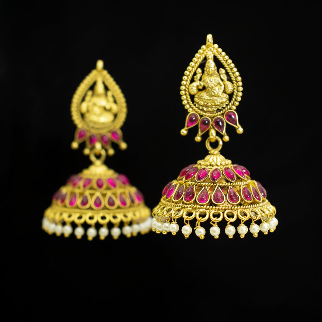 A pair of ornate gold-toned Indian jhumka earrings embellished with pink teardrop-shaped stones and small white bead drops, shown against a black background with a central Sahielli watermark.