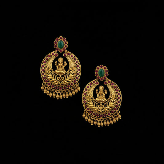 A pair of traditional Indian gold-plated jhumka earrings with red stone accents, green stone tops, and a central deity motif, displayed on a dark reflective surface.