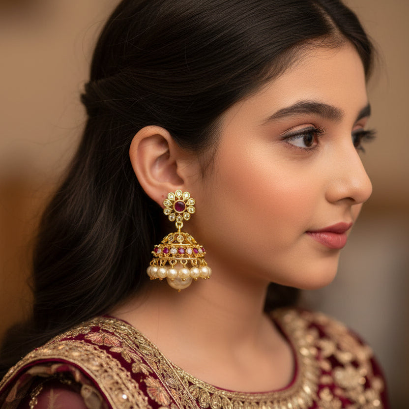 Gold earrings with pearls and red gemstones on a black stand, branded 'Saheli'.