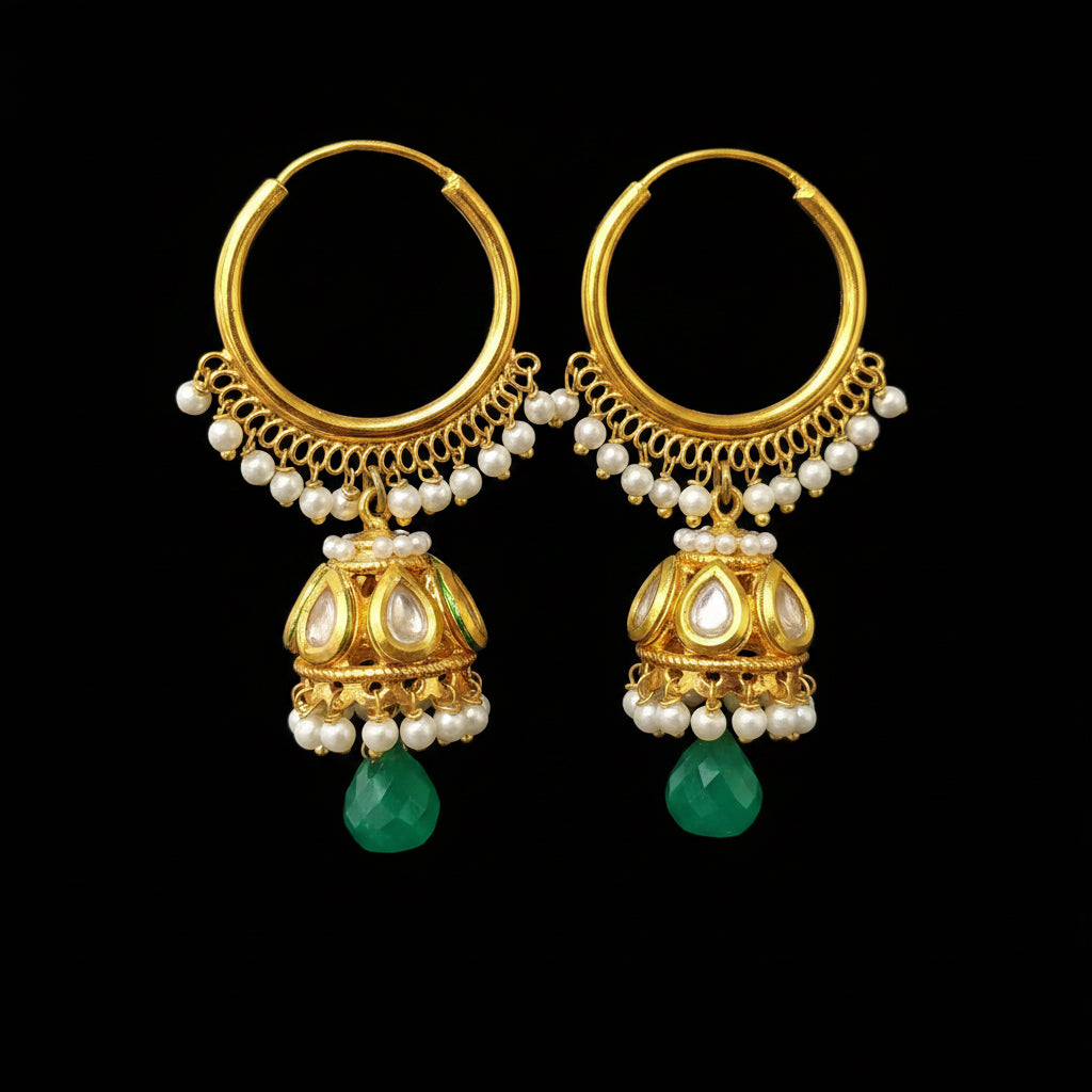 A pair of gold-toned traditional hoop earrings with dangling bell-shaped jhumka details, decorated with white pearl beads and green teardrop gemstones, displayed on a gray jewelry stand.