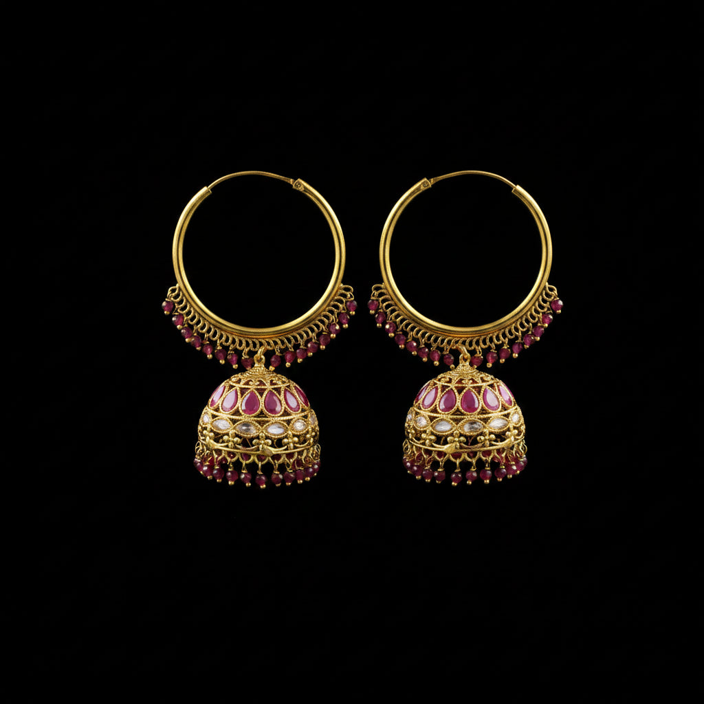 A pair of ornate gold-toned hoop earrings with dangling jhumka-style bells, decorated with pink gemstones and small red bead drops, displayed on a black stand against a gray background.
