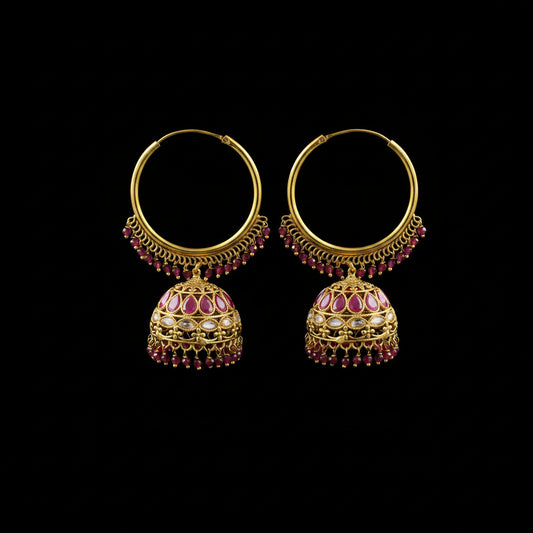 A pair of ornate gold-toned hoop earrings with dangling jhumka-style bells, decorated with pink gemstones and small red bead drops, displayed on a black stand against a gray background.