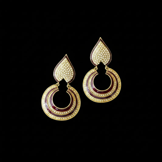 A pair of gold-toned teardrop-shaped earrings with concentric circular disc drops, decorated with gold, deep red enamel, and small clear rhinestone accents, displayed on a dark reflective surface with the SAHELI MY BEST FRIEND logo above.