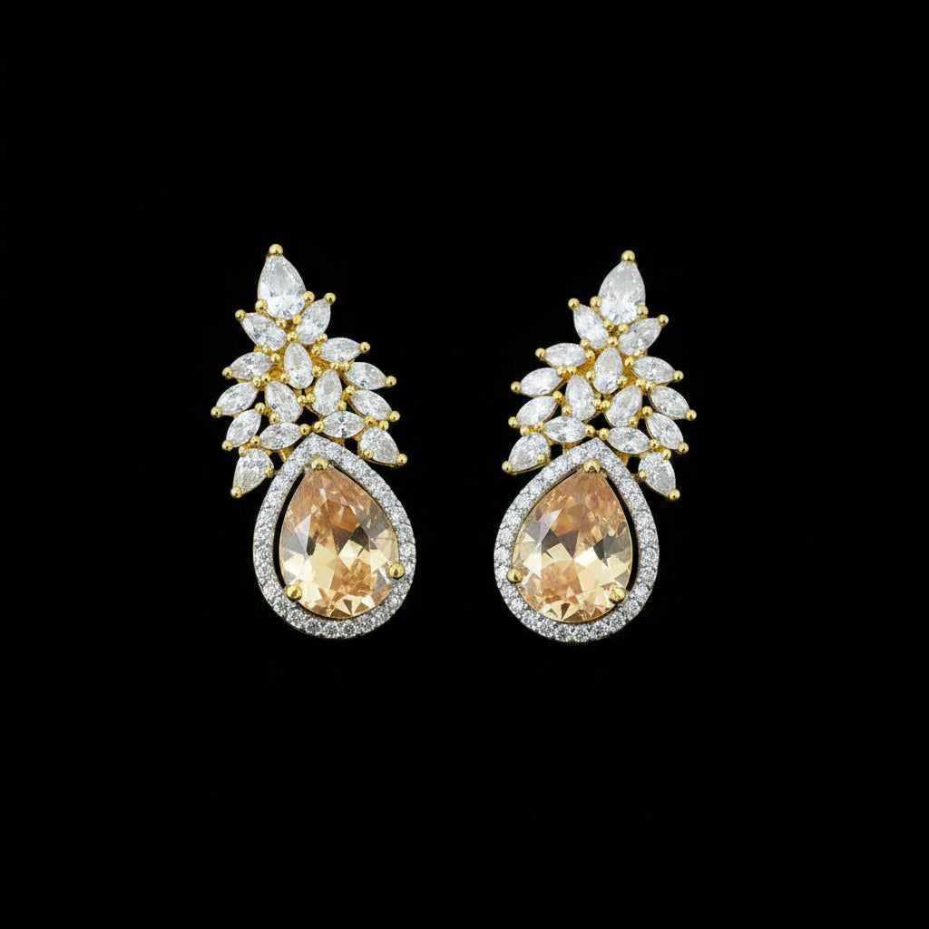 A pair of gold and silver stud earrings featuring pear-cut orange gemstones surrounded by clear crystals, displayed on a dark reflective surface with a Saheli logo in the top left corner.