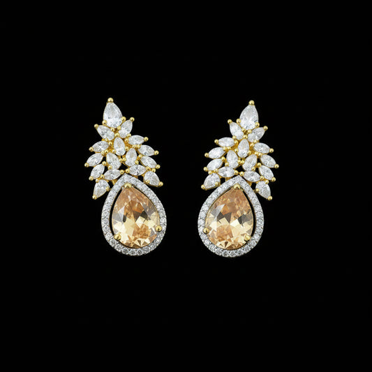 A pair of gold and silver stud earrings featuring pear-cut orange gemstones surrounded by clear crystals, displayed on a dark reflective surface with a Saheli logo in the top left corner.