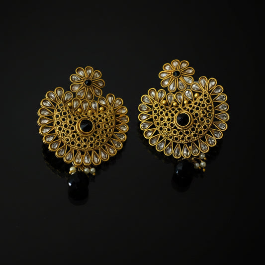 A pair of ornate gold-toned Indian jhumka earrings decorated with clear kundan stones and black bead accents, displayed on a reflective black surface. The brand logo 'SAHELI MY BESTFRIEND' is visible in the top left corner.